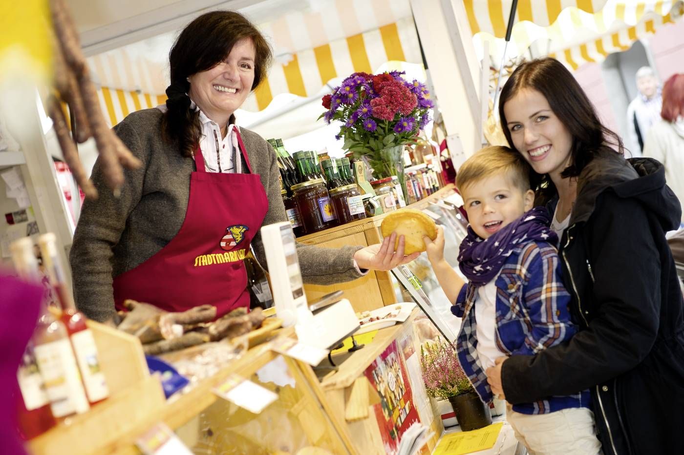 Eine junge Mutter kauft mit ihrem Sohn am Stadtmarkt einen Krapfen von einer sympathisch lächelnden Verkäuferin.